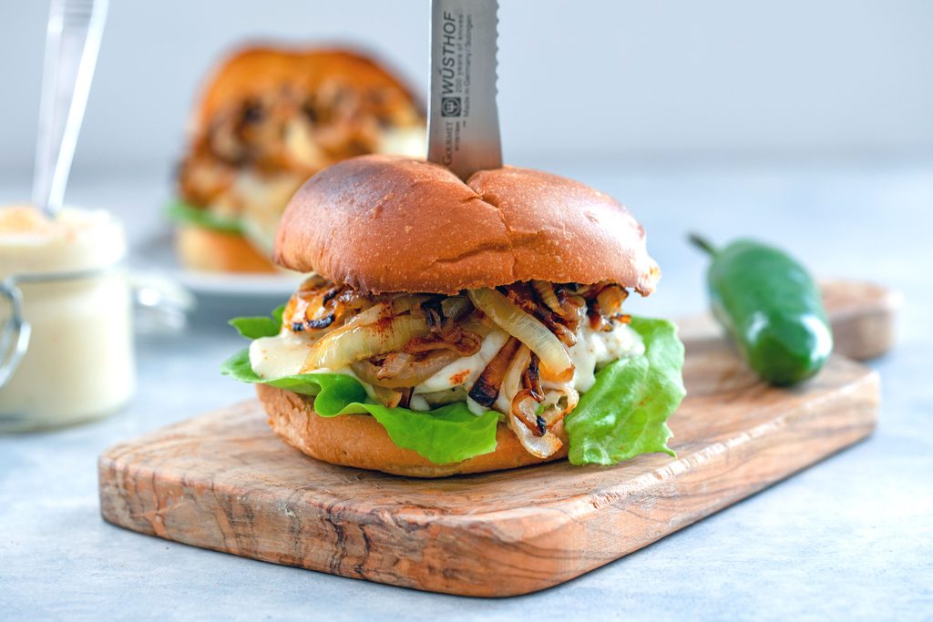 Landscape view of spicy cheesy chicken burger with cheese sauce and caramelized onions with a knife stuck in it on a small wooden cutting board with jar of cheese sauce, second burger, and jalapeño pepper in background