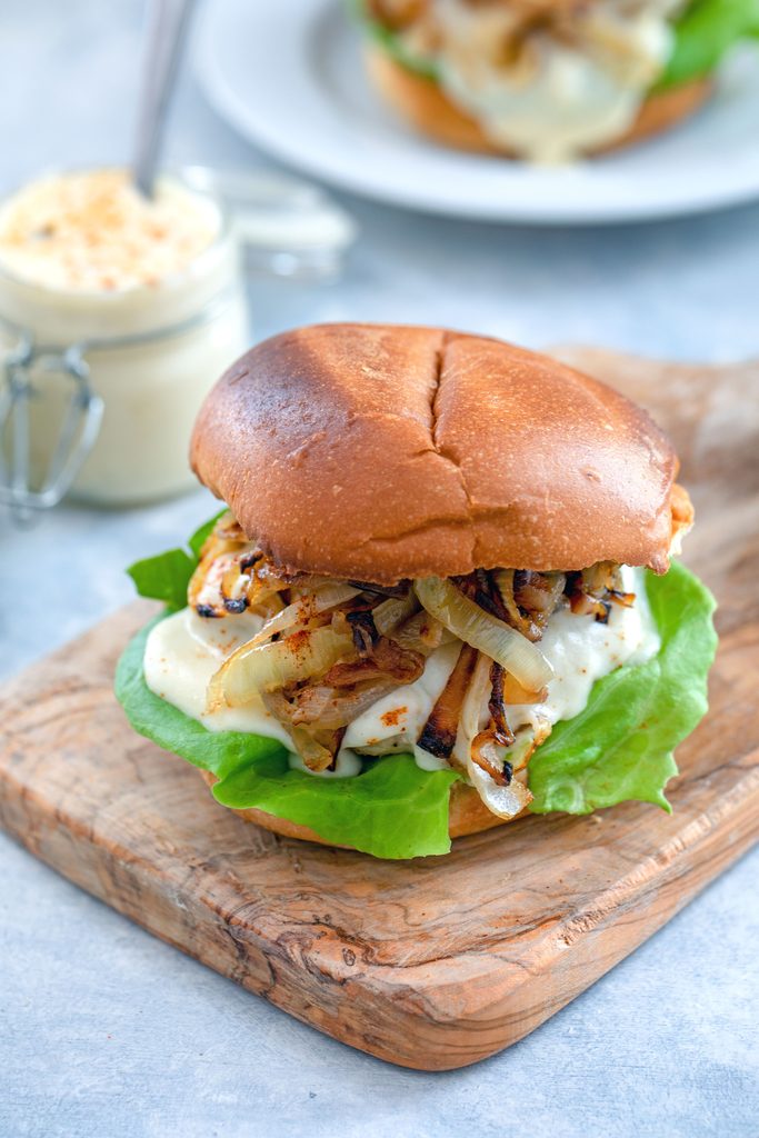 Head-on view of spicy cheesy chicken burger with caramelized onions on a small wooden cutting board with jar of cheese sauce and second burger in the background