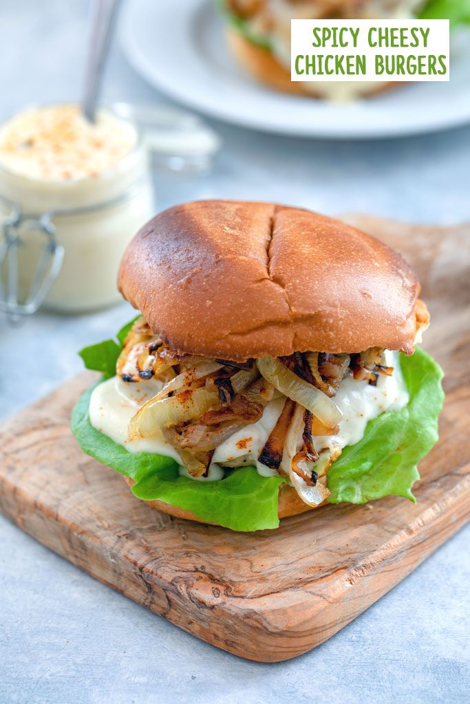 Head-on view of spicy cheesy chicken burger with caramelized onions on a small wooden cutting board with jar of cheese sauce and second burger in the background with recipe title at top