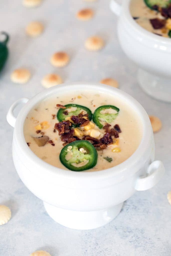 Overhead view of a white bowl of spicy clam chowder with corn topped with bacon and jalapeños and surrounded by oyster crackers