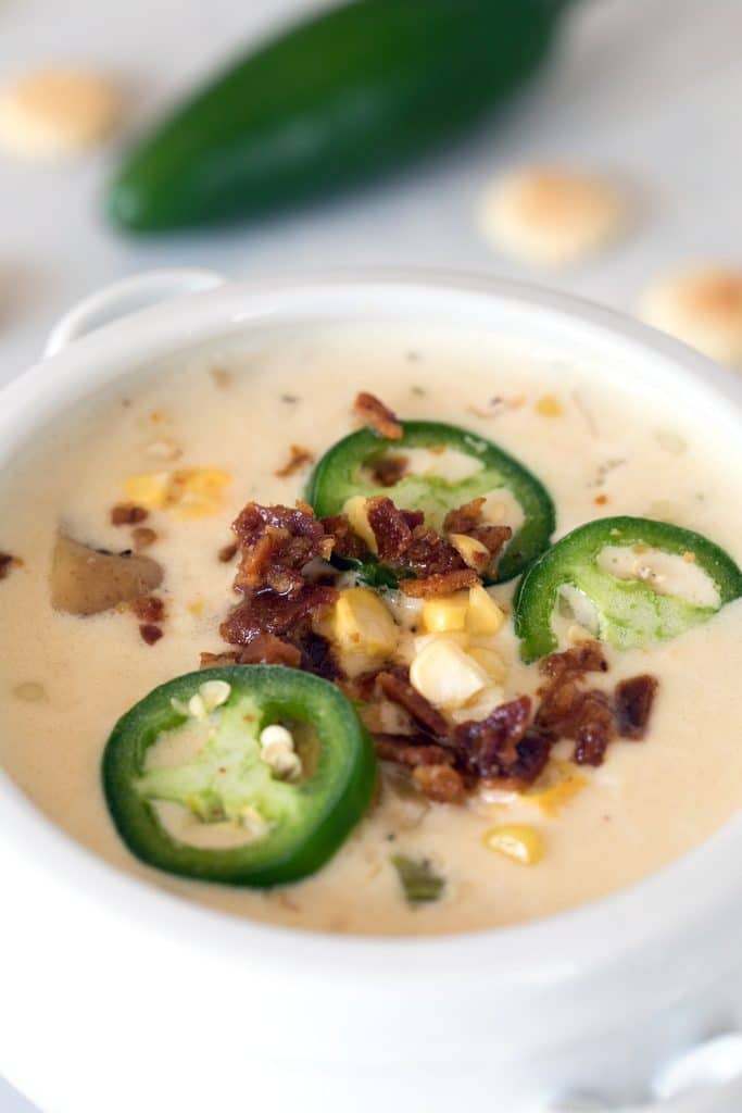 Closeup view of a white bowl of spicy clam chowder with corn topped with jalapeño slices and bacon crumbles with oyster crackers and jalapeño in background