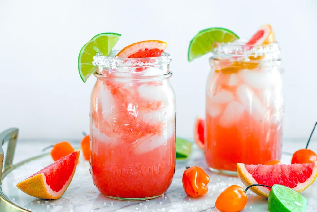 Landscape view of two spicy grapefruit margaritas in mason jars on marble tray, surrounded by grapefruit wedges, lime wedges, and habanero peppers