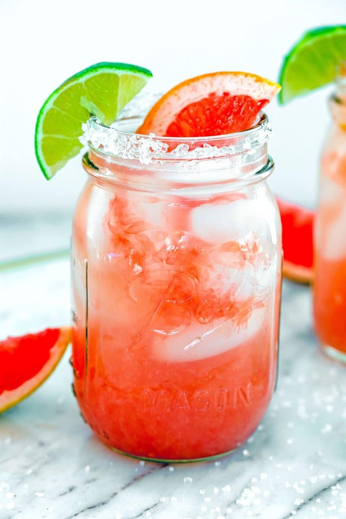 Head-on close-up view of spicy grapefruit margarita with grapefruit and lime garnish with salt on marble surface