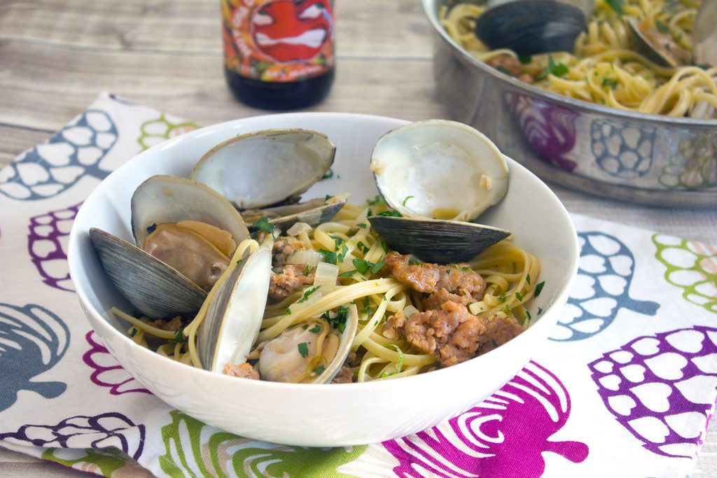 Landscape head-on view of a big white bowl of spicy lime linguine with clams and sausage on a colorful tea towel with bottle of cider and pan of linguine and clams in the background