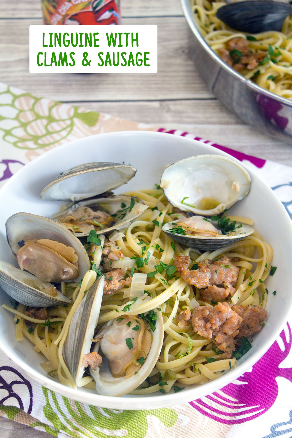 Overhead view of a big bowl of linguine with clams and sausage with a hard cider in the background and recipe title at top.