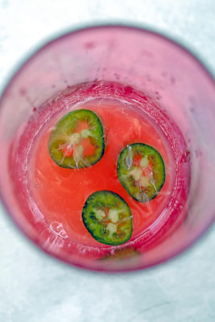 Overhead view of sliced jalapeños, lime juice, and whiskey in pink cocktail shaker