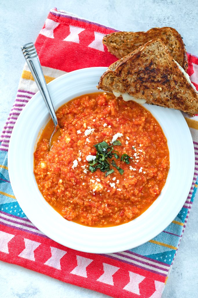 Overhead view of spicy roasted tomato soup with quinoa and feta in a white bowl with a halved grilled cheese sandwich on the side and on a colorful dish towel