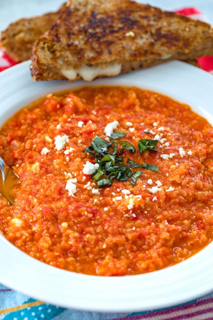 Closeup view of spicy roasted tomato soup with quinoa and feta topped with basil and feta in a white bowl with a halved grilled cheese sandwich on the side