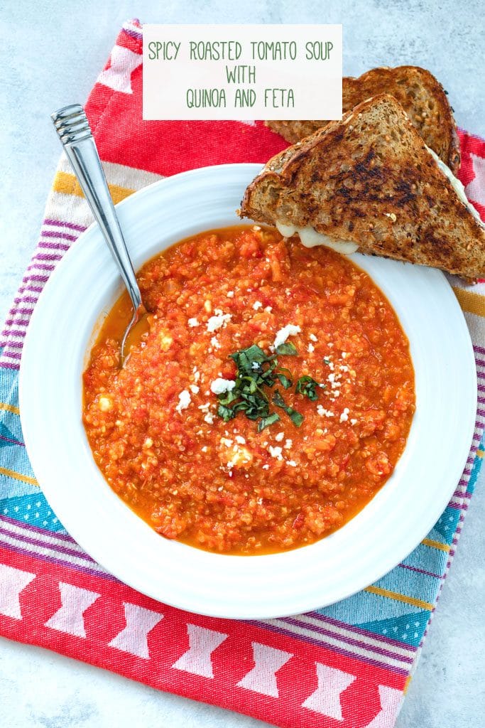 Overhead view of spicy roasted tomato soup with quinoa and feta in a white bowl with a halved grilled cheese sandwich on the side and on a colorful dish towel with "Spicy Roasted Tomato Soup with Quinoa and Feta" text at top