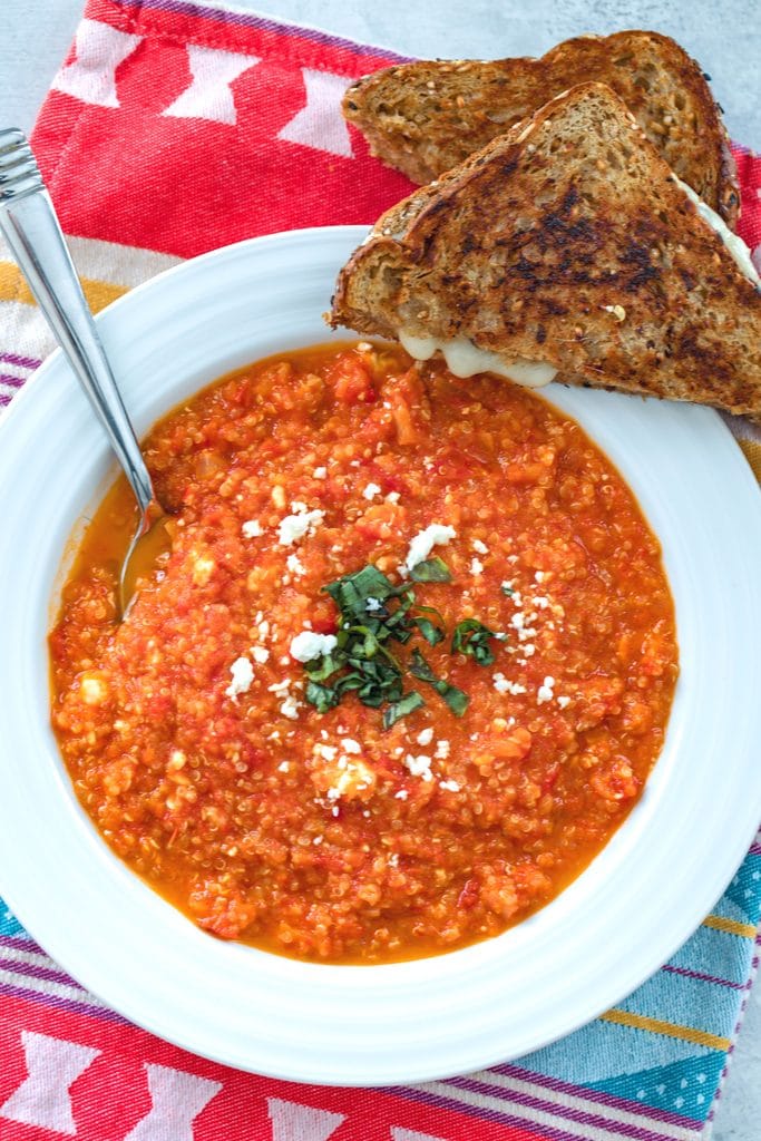 Overhead closeup view of white bowl with spicy roasted tomato soup with feta and quinoa and a halved grilled cheese sandwich on the side on a colorful dish towel