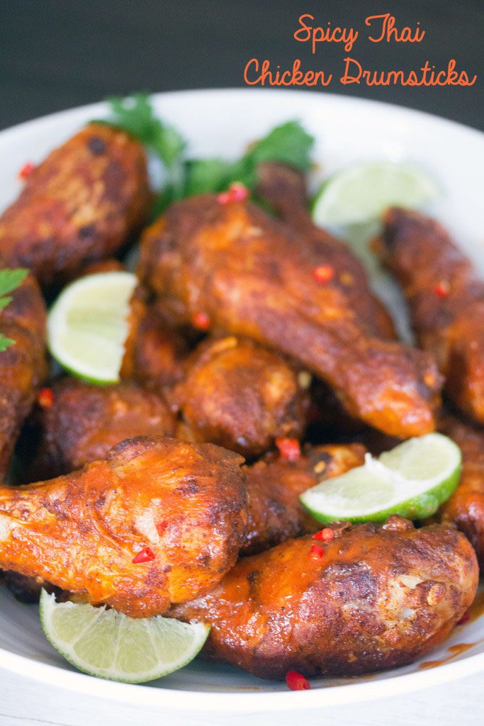 Head-on view of a white serving bowl filled with Spicy Thai Chicken Drumsticks with lime wedges and parsley with recipe title at the top of the image