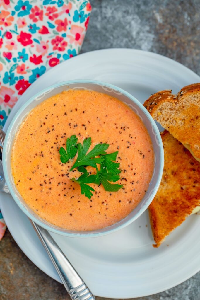 Overhead view of a bowl of spicy tomato soup with parsley garnish and grilled cheese sandwich on the side