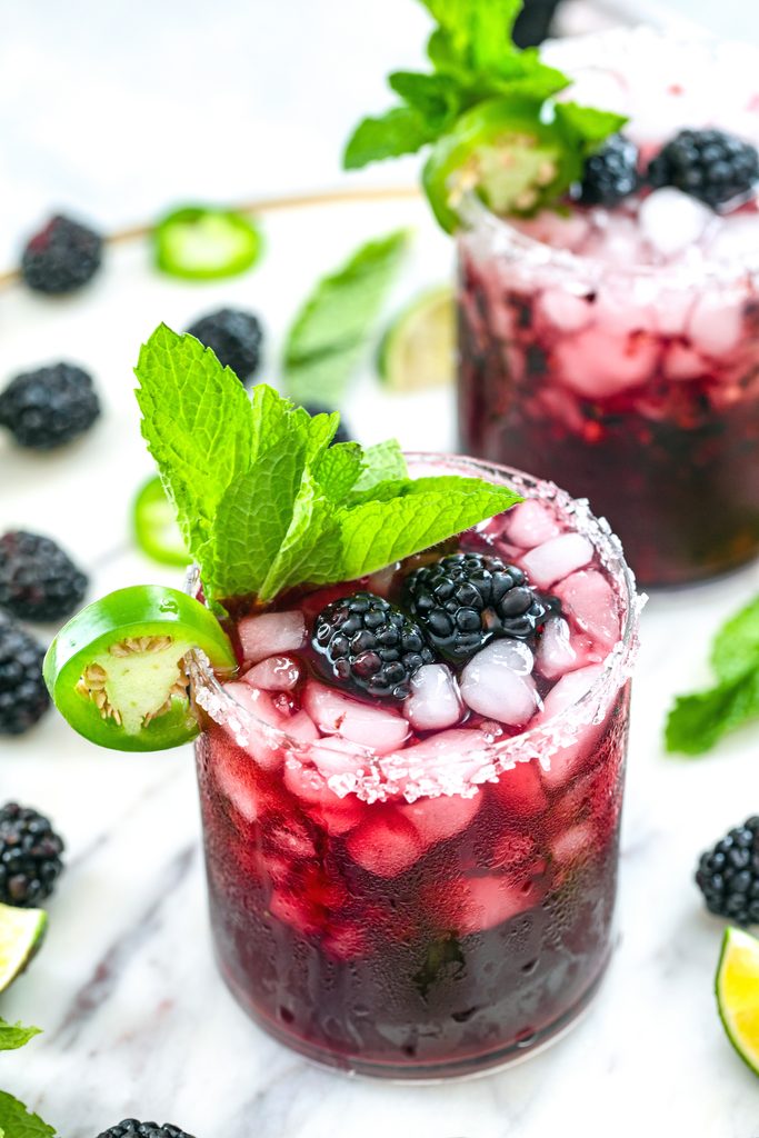 Overhead view of a spicy vodka blackberry smash cocktail on a marble surface with second cocktail and blackberries and jalapeño slices scattered around