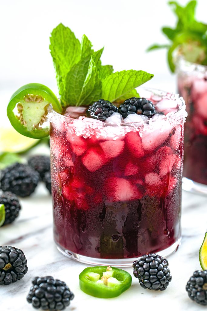 Head-on closeup view of a spicy vodka blackberry smash cocktail with sugared rim and mint, blackberry, and jalapeño garnish with blackberries and jalapeño slices all around