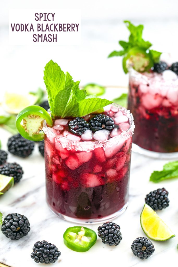 Head-on view of a spicy vodka blackberry smash on a marble surface with second cocktail, blackberries, jalapeño slices, and lime wedges in the background and recipe title at top