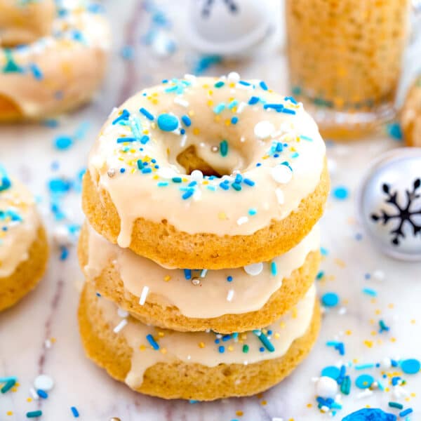 Head-on view of a stack of three spiked eggnog doughnuts with more doughnuts, glass of eggnog, jingle bells, and blue and white sprinkles in background