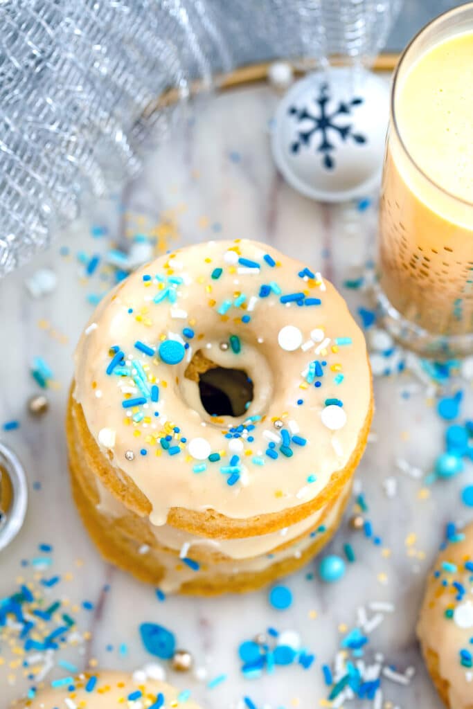 Bird's eye view of a stack of eggnog doughnuts on a marble tray with glass of eggnog in background and sprinkles and jingle bells all around