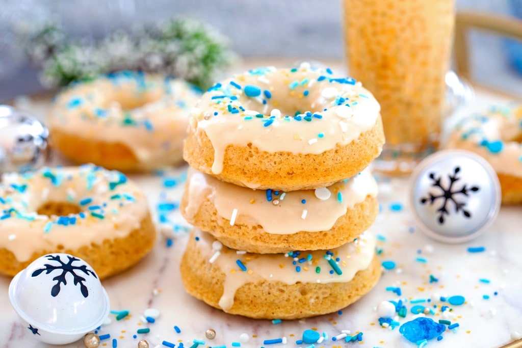 Landscape head-on view of a stack of three eggnog doughnuts with more donuts, sprinkles, jingle bells, and glass of eggnog in background