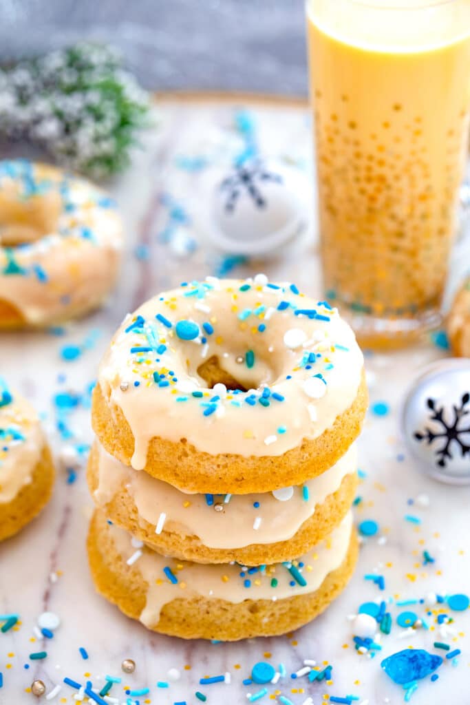 Head-on view of a stack of three spiked eggnog doughnuts with more doughnuts, glass of eggnog, jingle bells, and blue and white sprinkles in background