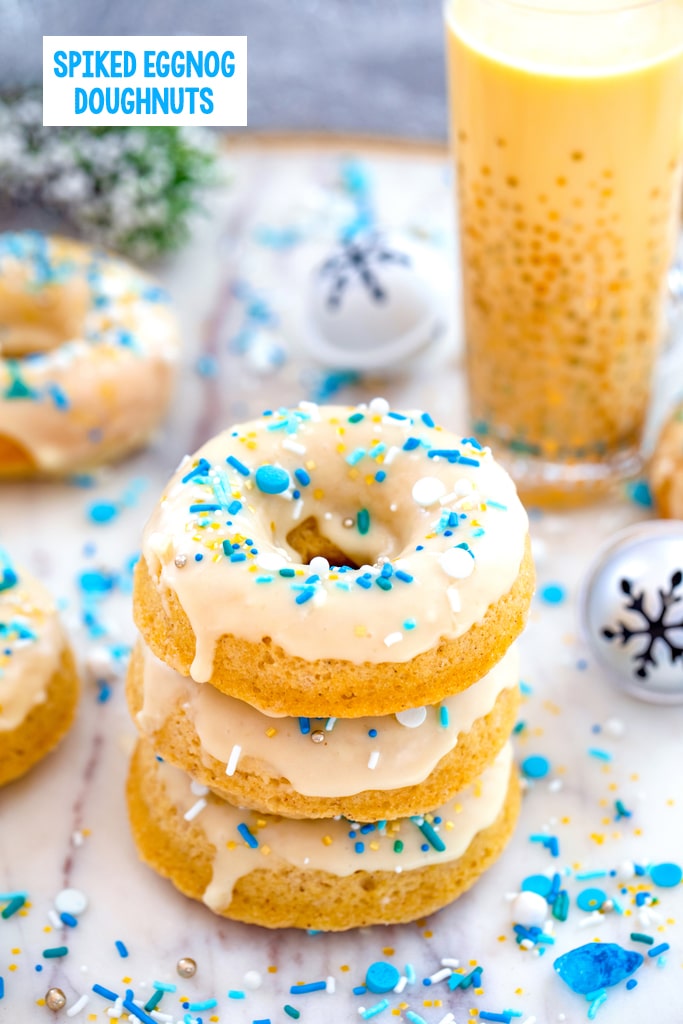 Head-on view of a stack of three spiked eggnog doughnuts with more doughnuts, glass of eggnog, jingle bells, and blue and white sprinkles in background with recipe title at top