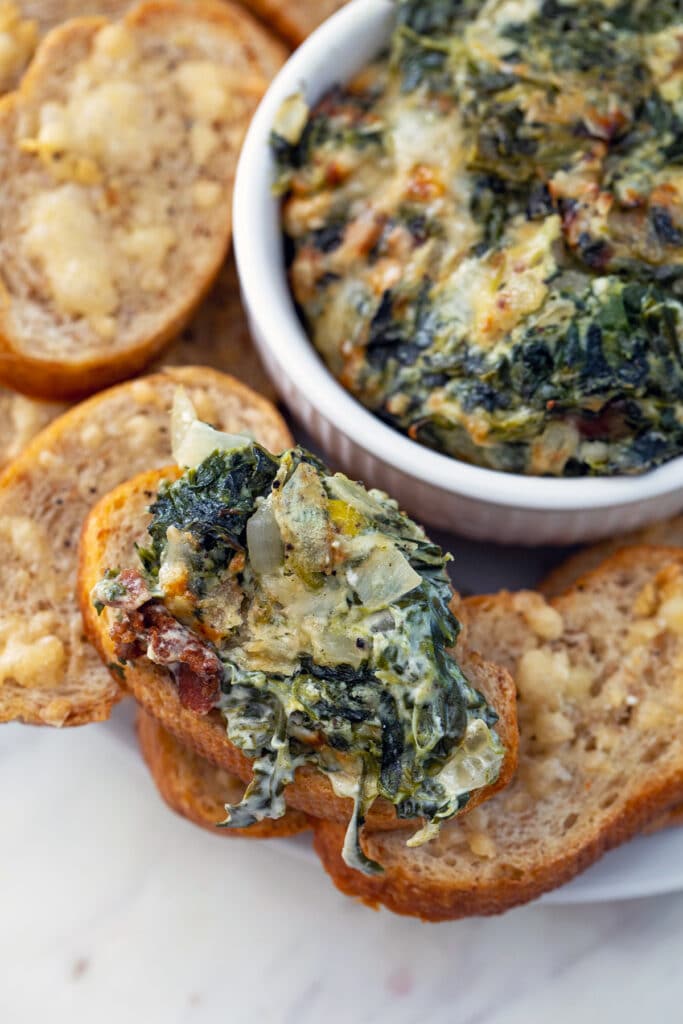Overhead view of a garlic toast topped with spinach dip with onion and bacon with more garlic toasts and bowl with dip