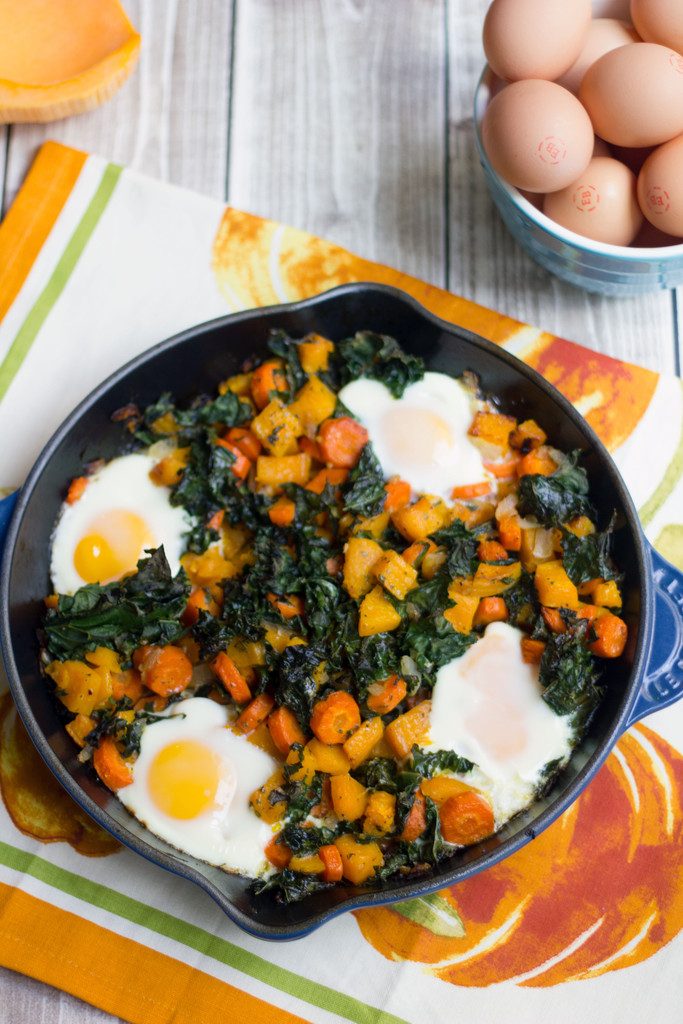 Bird's eye view of a cast iron skillet with squash hash with kale and eggs with half a butternut squash and bowl of eggs in the background