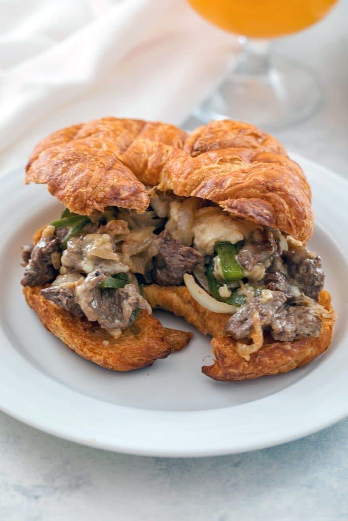 Steak and cheese croissant with steak, green peppers, onions, and white cheese on a white plate with a white napkin and glass of beer in the background