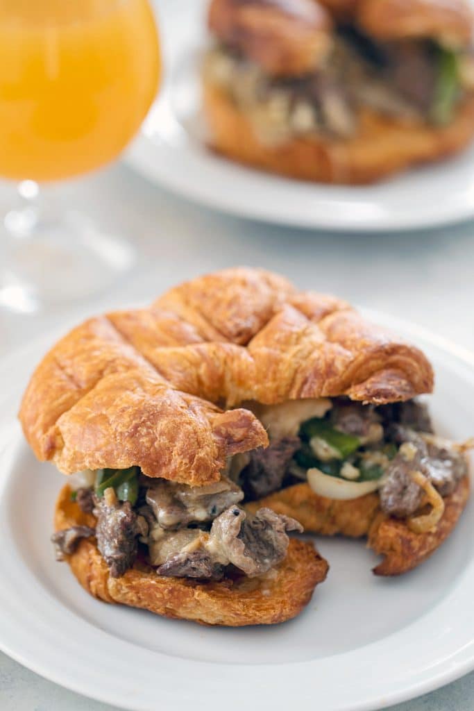 Head-on view of steak and cheese croissant on a white plate with a second sandwich in the background and a glass of beer