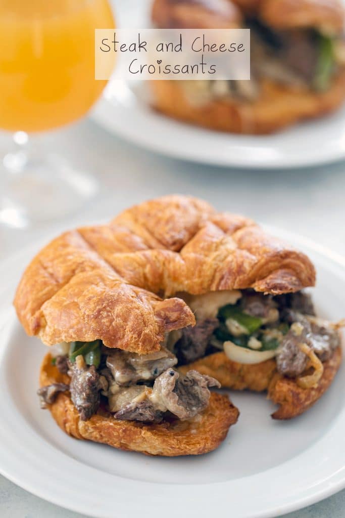 Head-on view of steak and cheese croissant on a white plate with a second sandwich in the background and a glass of beer with "Steak and Cheese Croissants" text at the top