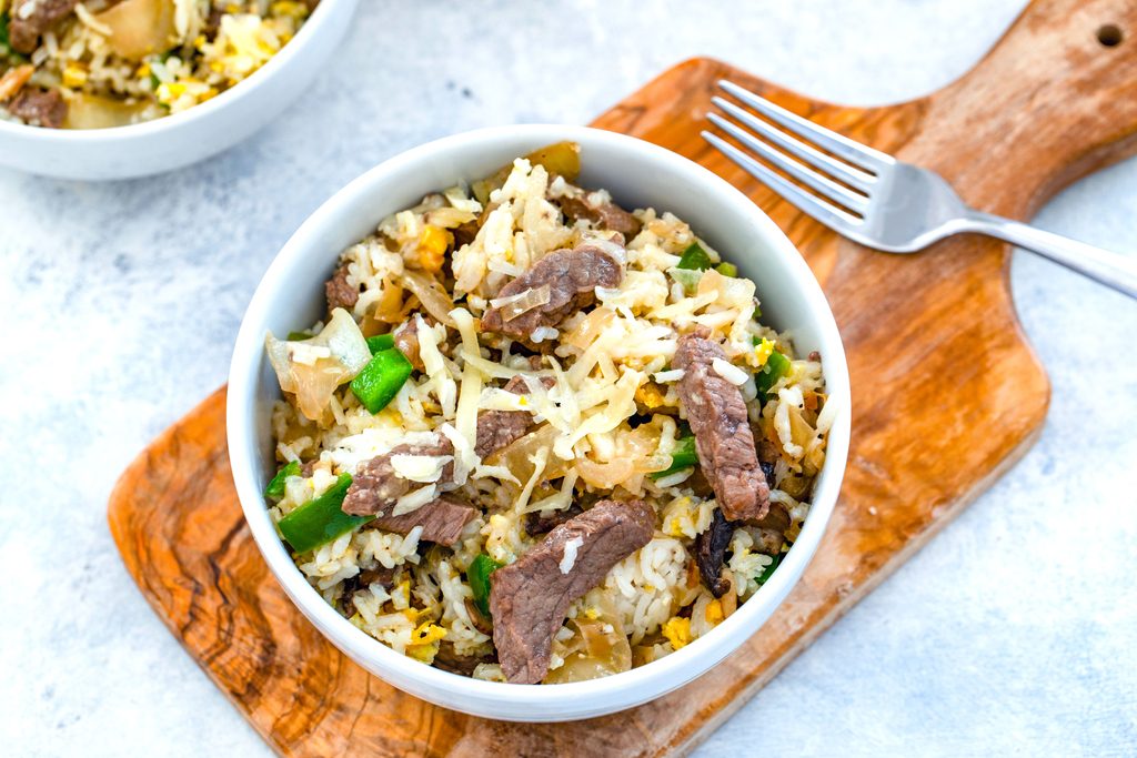 Landscape overhead view of bowl of steak and cheese fried rice on a wooden board with fork and second bowl in the background