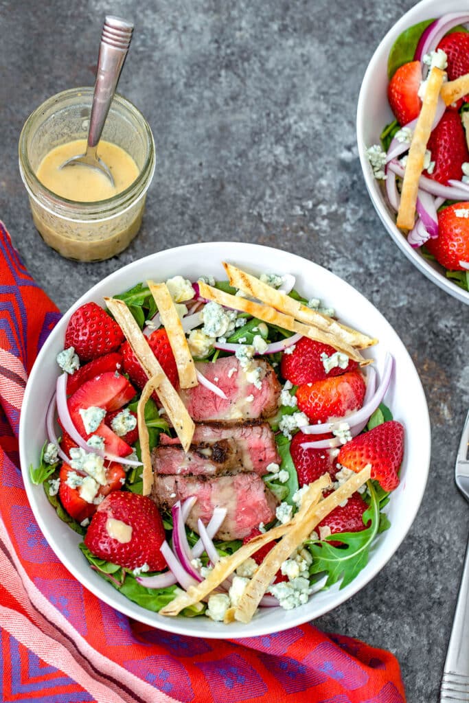Bird's eye view of steak and strawberry salad in white bowl with greens, blue cheese, red onion, and tortilla strips.
