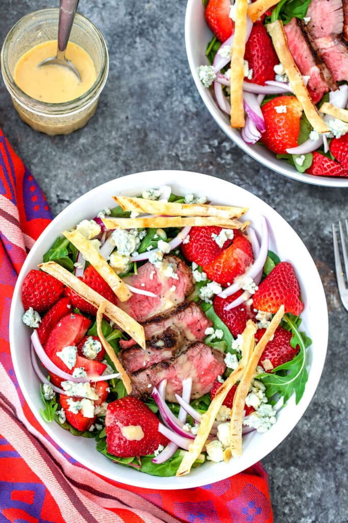 Overhead view of a steak and strawberry salad in a bowl with greens, blue cheese, red onion, and tortilla strips with jar of lemon dressing, second bowl of salad in background, and recipe title at top..