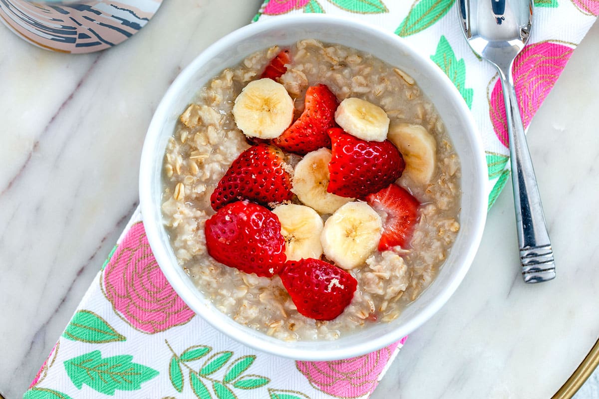 Landscape overhead view of strawberry banana oatmeal on a pink and green towel on a marble surface with a spoon in the background.