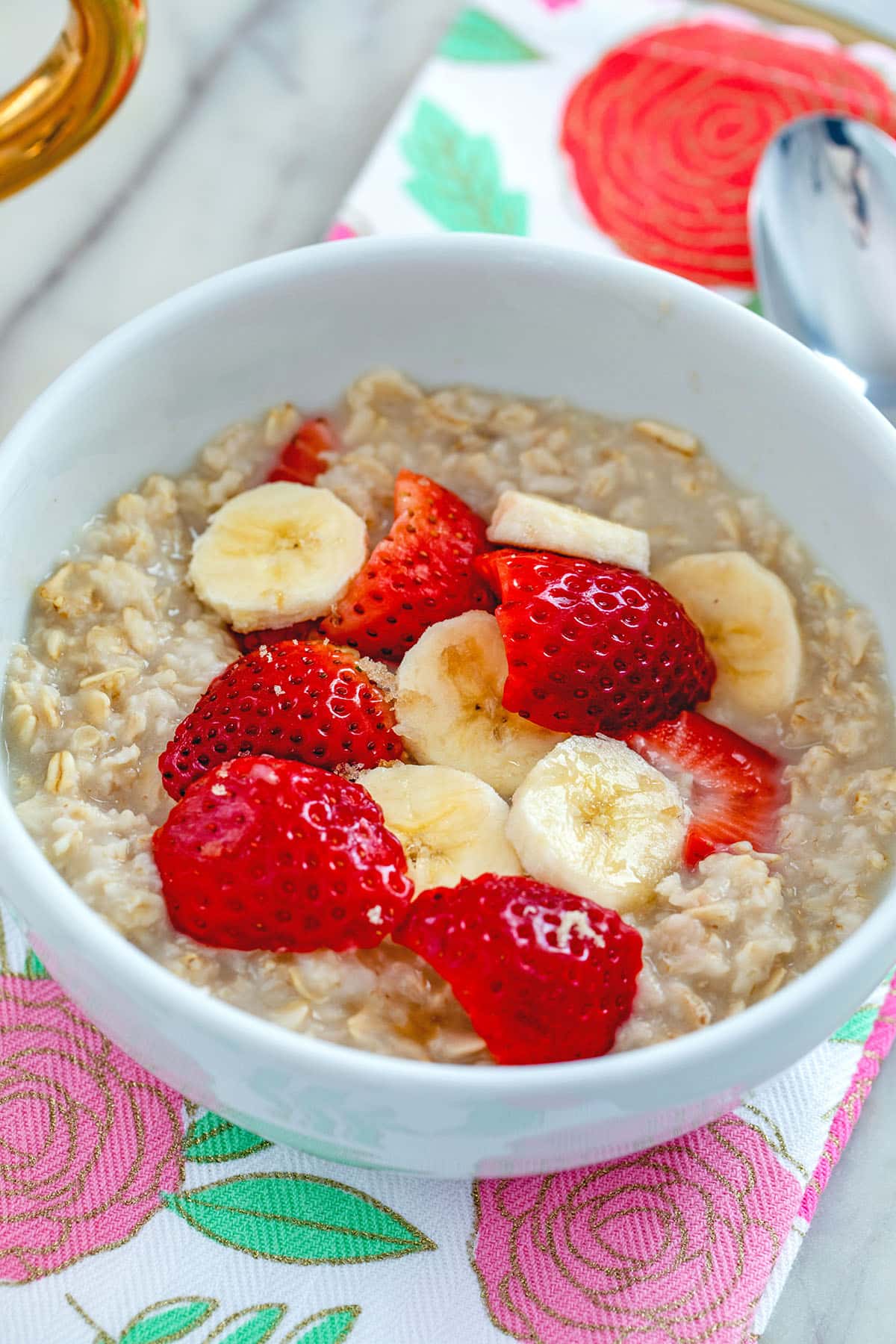 Head-on view of a white bowl of strawberry banana oatmeal on a pink and red flowered towel with spoon.