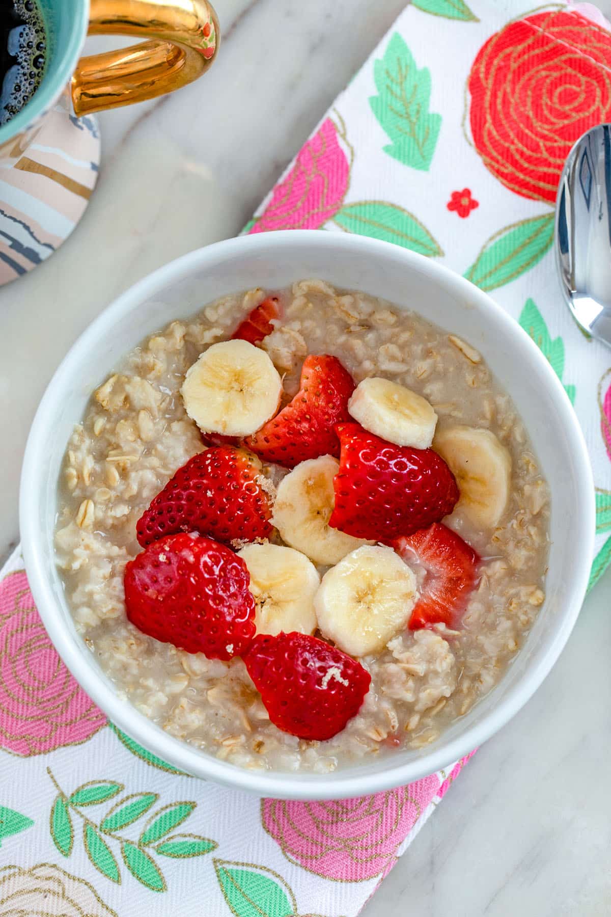 Overhead view of strawberry banana oatmeal in a white bowl on a pink and red flower towel with spoon and cup of coffee in the background.