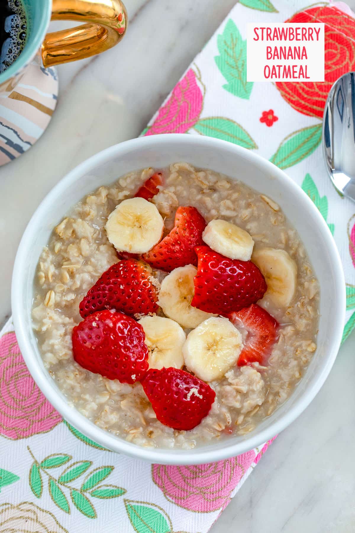 Overhead view of strawberry banana oatmeal in a white bowl on a pink and red flower towel with spoon and cup of coffee in the background and recipe title at top.