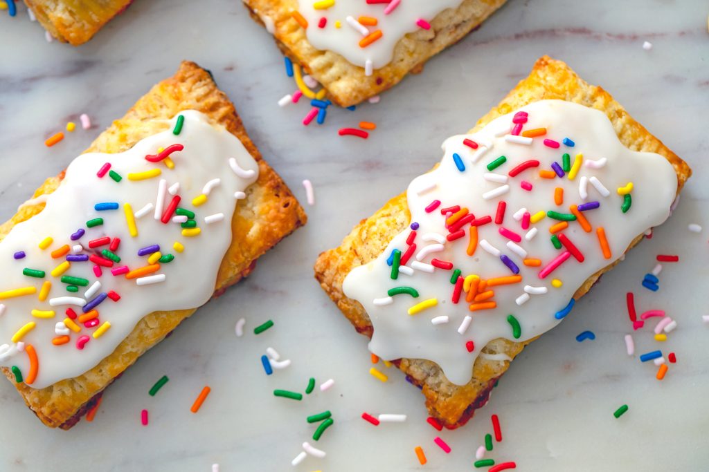 Landscape view of strawberry breakfast tarts laying on marble surface and covered in white icing and rainbow sprinkles with more sprinkles scattered around