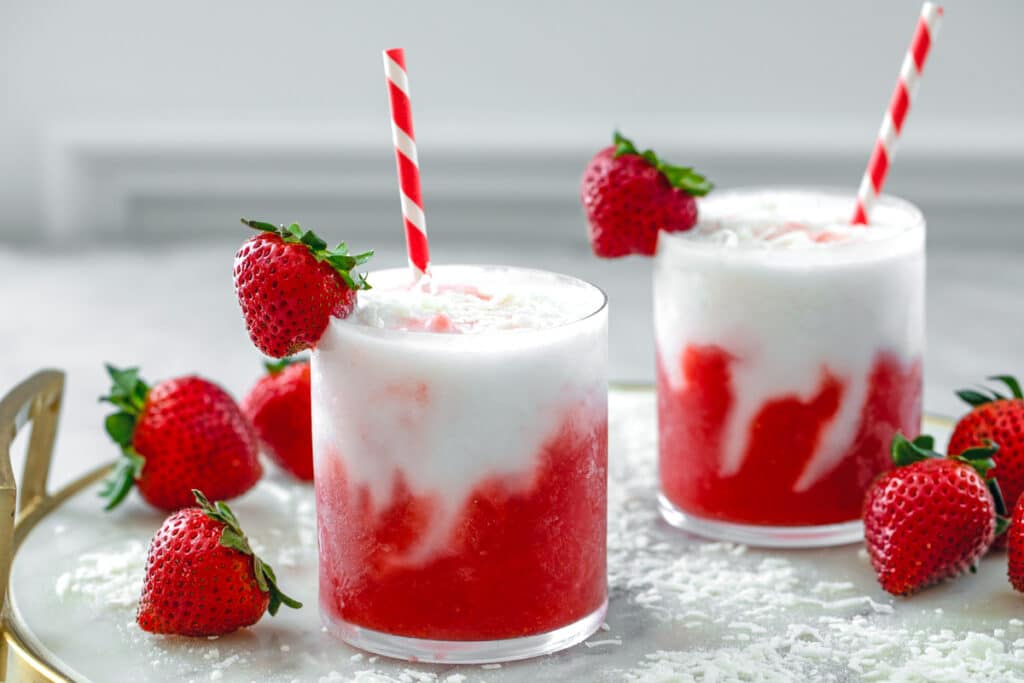 Landscape head-on view of two strawberry coconut daiquiris on a marble and gold tray with shredded coconut and strawberries all around