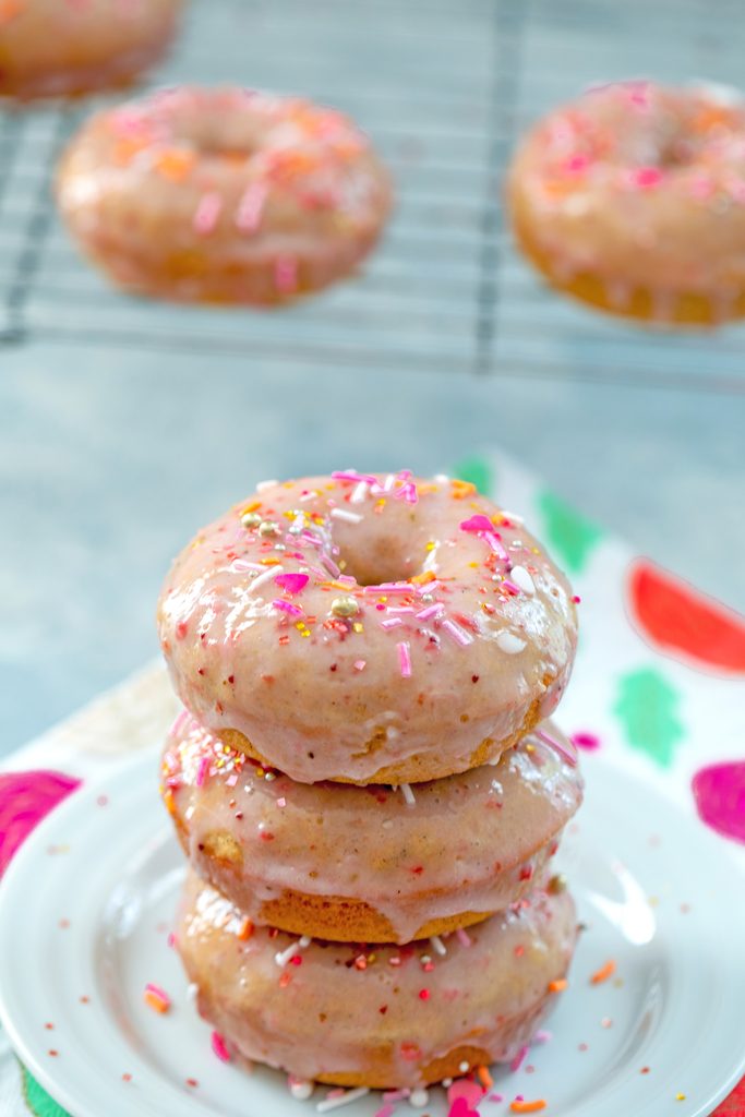 Head-on view of a stack of three strawberry frosted doughnuts covered in sprinkles on a white plate with a metal rack with more doughnuts on it in the backgrouond