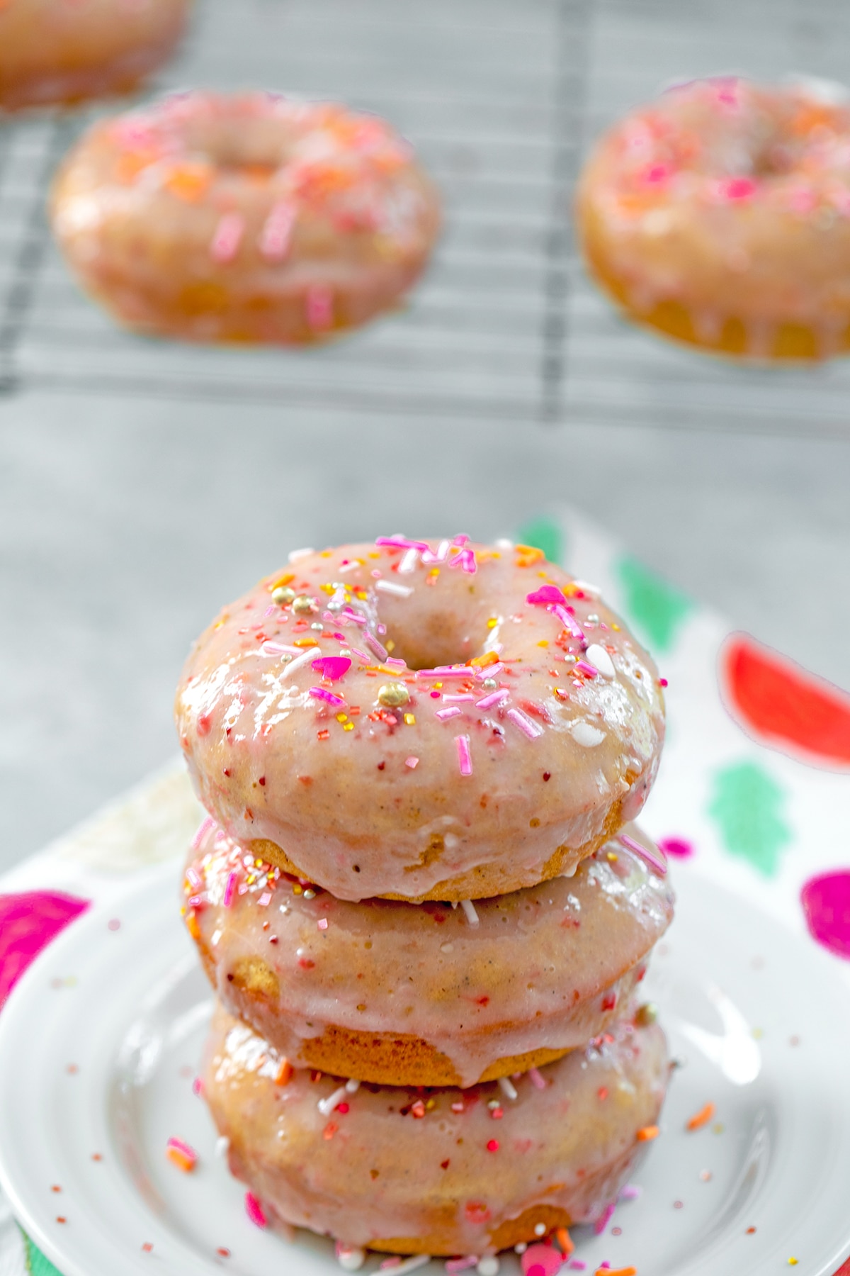 Head-on view of a stack of three strawberry frosted donuts covered in sprinkles on a white plate with a metal rack with more donuts on it in the background.