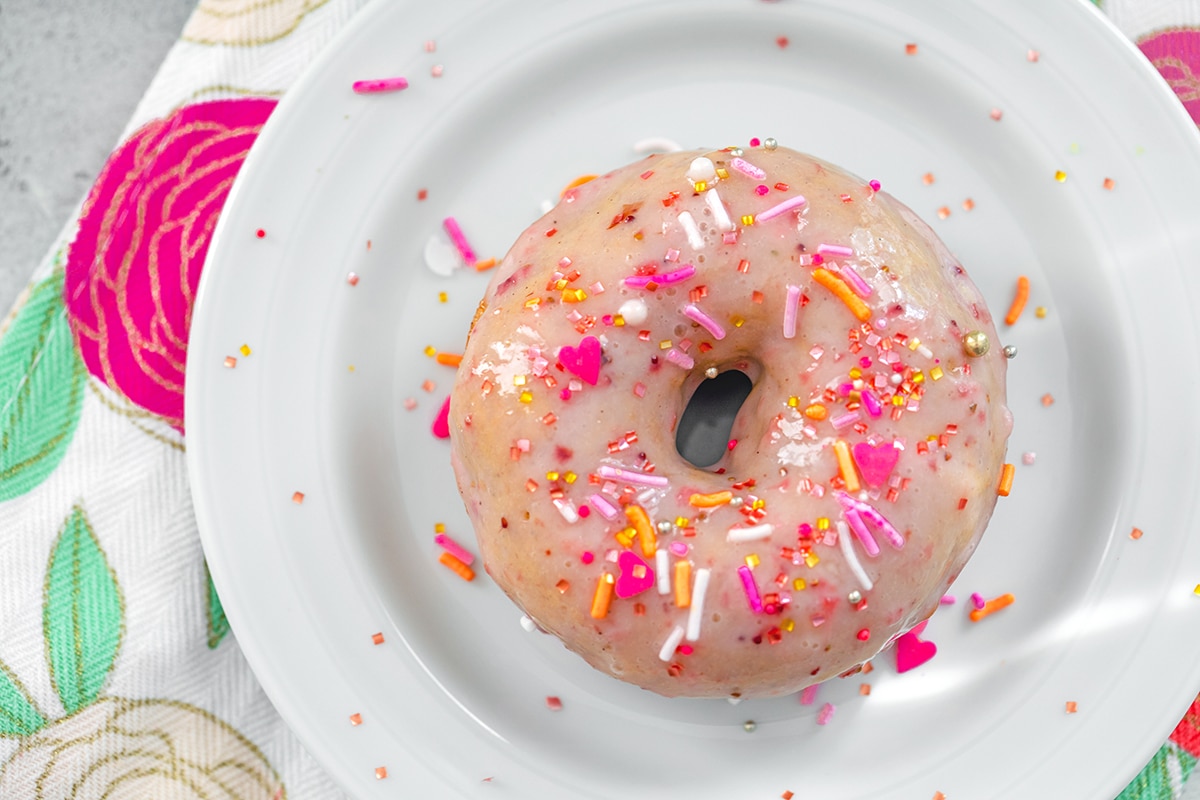 Landscape overhead view of strawberry frosted doughnut covered in pink and orange sprinkles on a white plate over a flowered towel.