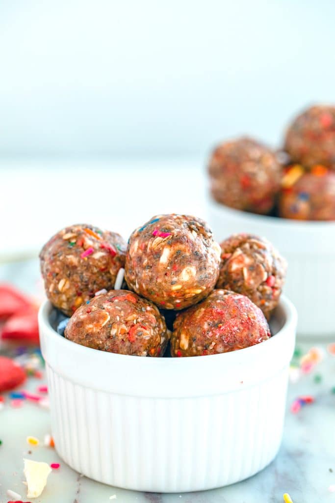 Head-on closeup view of strawberry funfetti energy bites in a small white bowl with second bowl in background and white chocolate, sprinkles, and dried strawberries all around