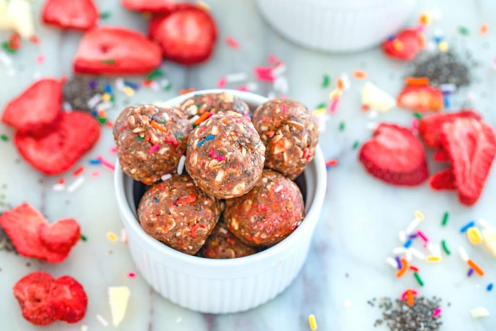 Landscape overhead view of strawberry funfetti energy bites piled into white bowl on a marble surface with dried strawberries, white chocolate, sprinkles, and chia seeds all around