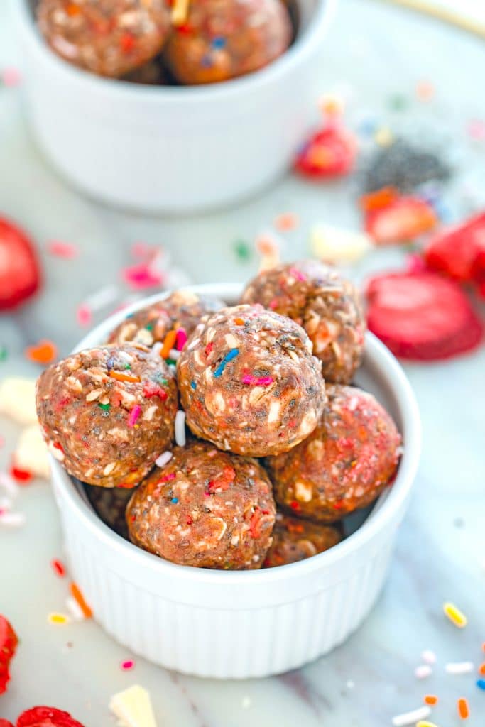 Overhead view of a white bowl filled with strawberry funfetti energy bites with a second bowl in the background, strawberries, sprinkles, chia seeds, and white chocolate sprinkled around