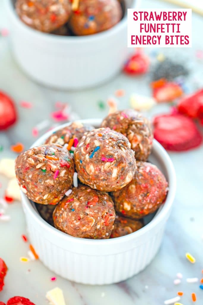 Overhead view of a white bowl filled with strawberry funfetti energy bites with a second bowl in the background, strawberries, sprinkles, chia seeds, and white chocolate sprinkled around and recipe title at top of image