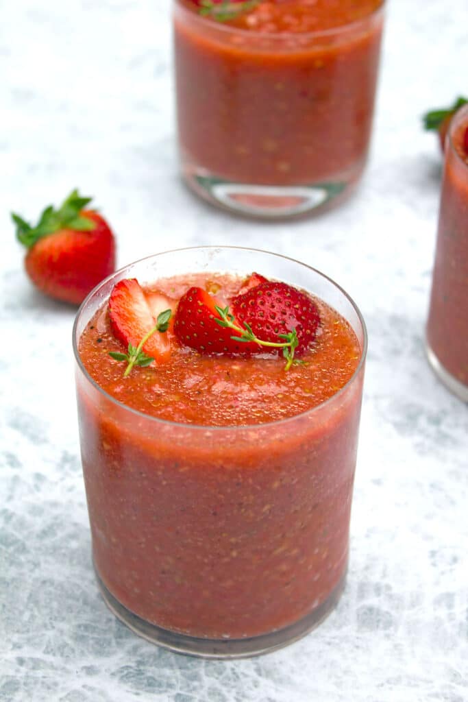 Overhead view of a clear glass of red strawberry gazpacho with sliced strawberry and thyme garnish with more glasses of gazpacho and a strawberry in the background