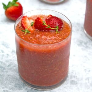 Closeup view of a cup of strawberry gazpacho with sliced strawberries and thyme garnish and strawberries in background
