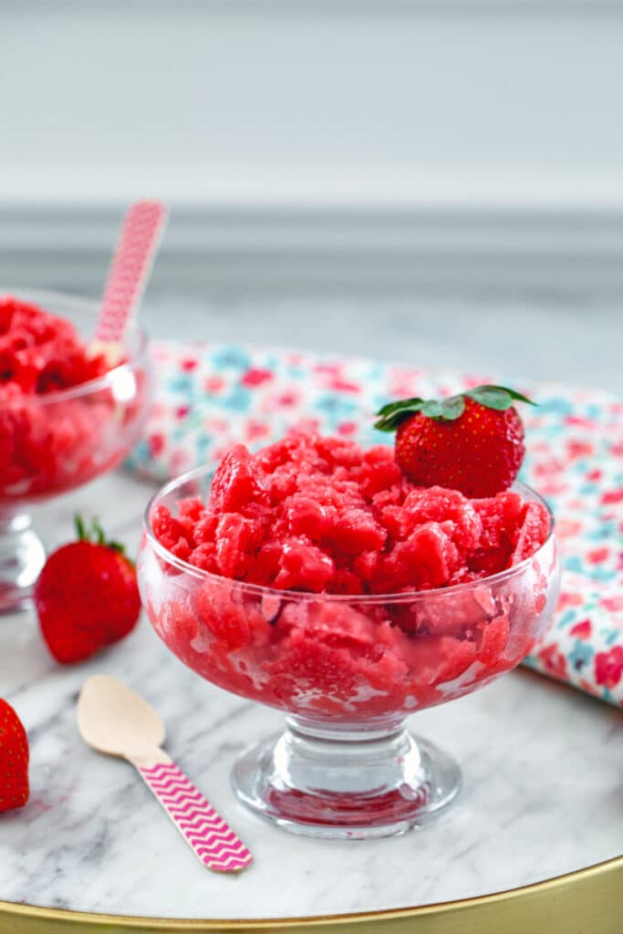 Head-on view of glass dishes of strawberry granita with fresh strawberries and a wooden spoon on a marble tray