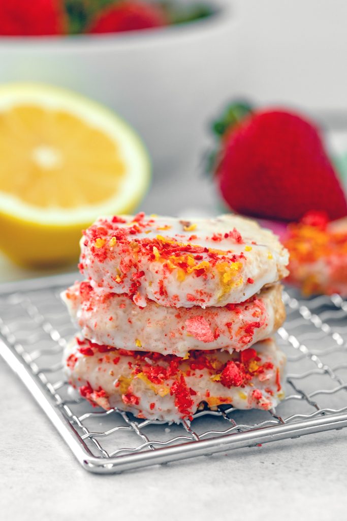 Head-on view of three strawberry lemonade shortbread cookies stacking on a baking rack with half a lemon and strawberries in background