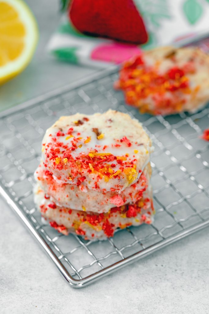 Overhead view of a stack of strawberry lemonade shortbread cookies on a metal rack with more cookies, a lemon half, and a strawberry in the background